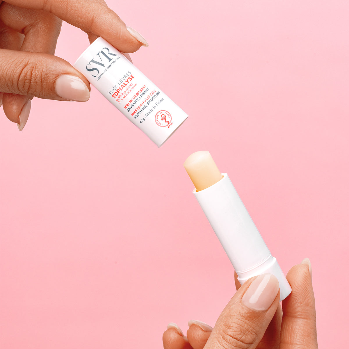 Bálsamo labial SVR Topialyse en envase blanco cilíndrico con tapa abierta, sostenido por manos sobre fondo rosa.