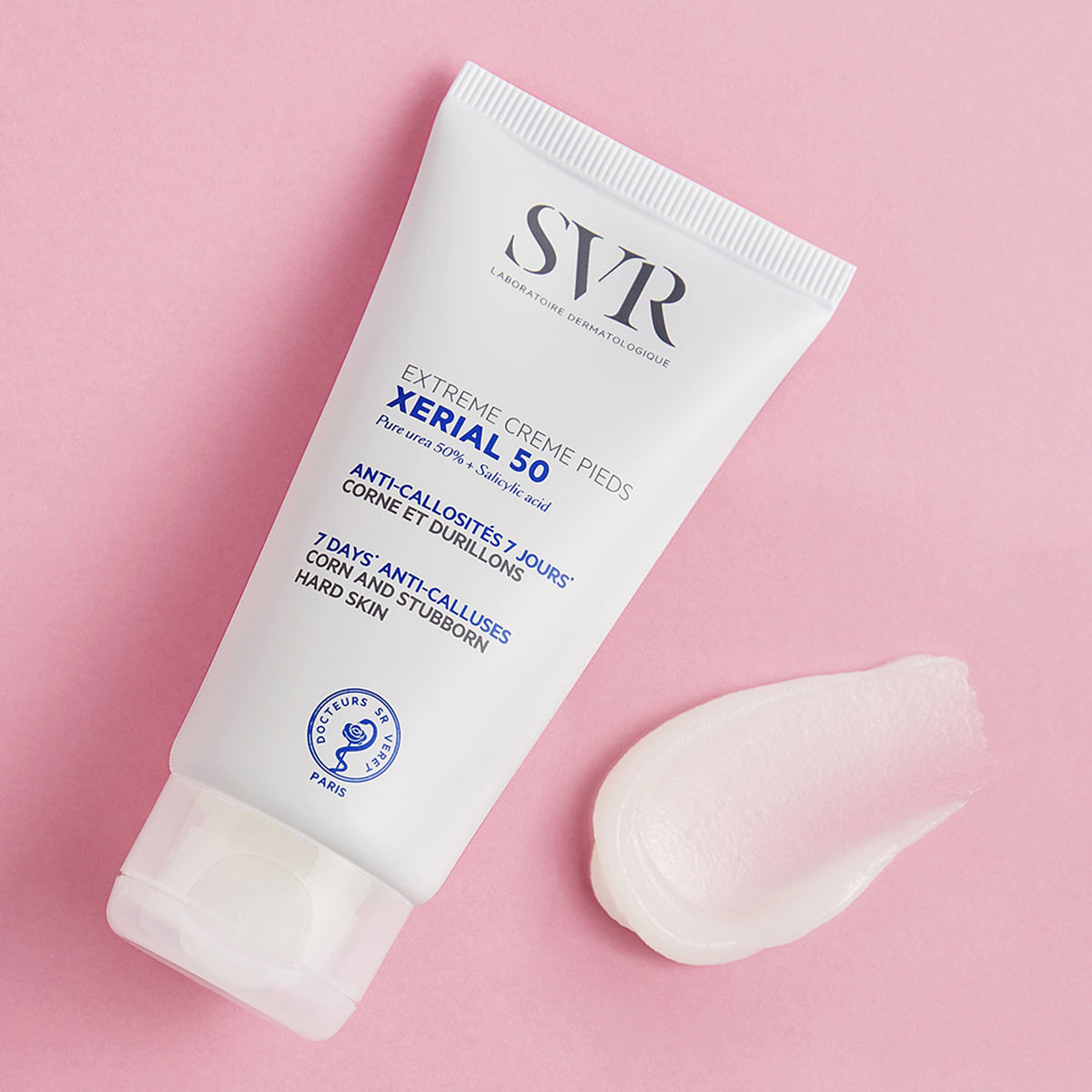 Tubo blanco de crema SVR XERIAL 50 para pies con muestra de textura sobre fondo rosa pastel.