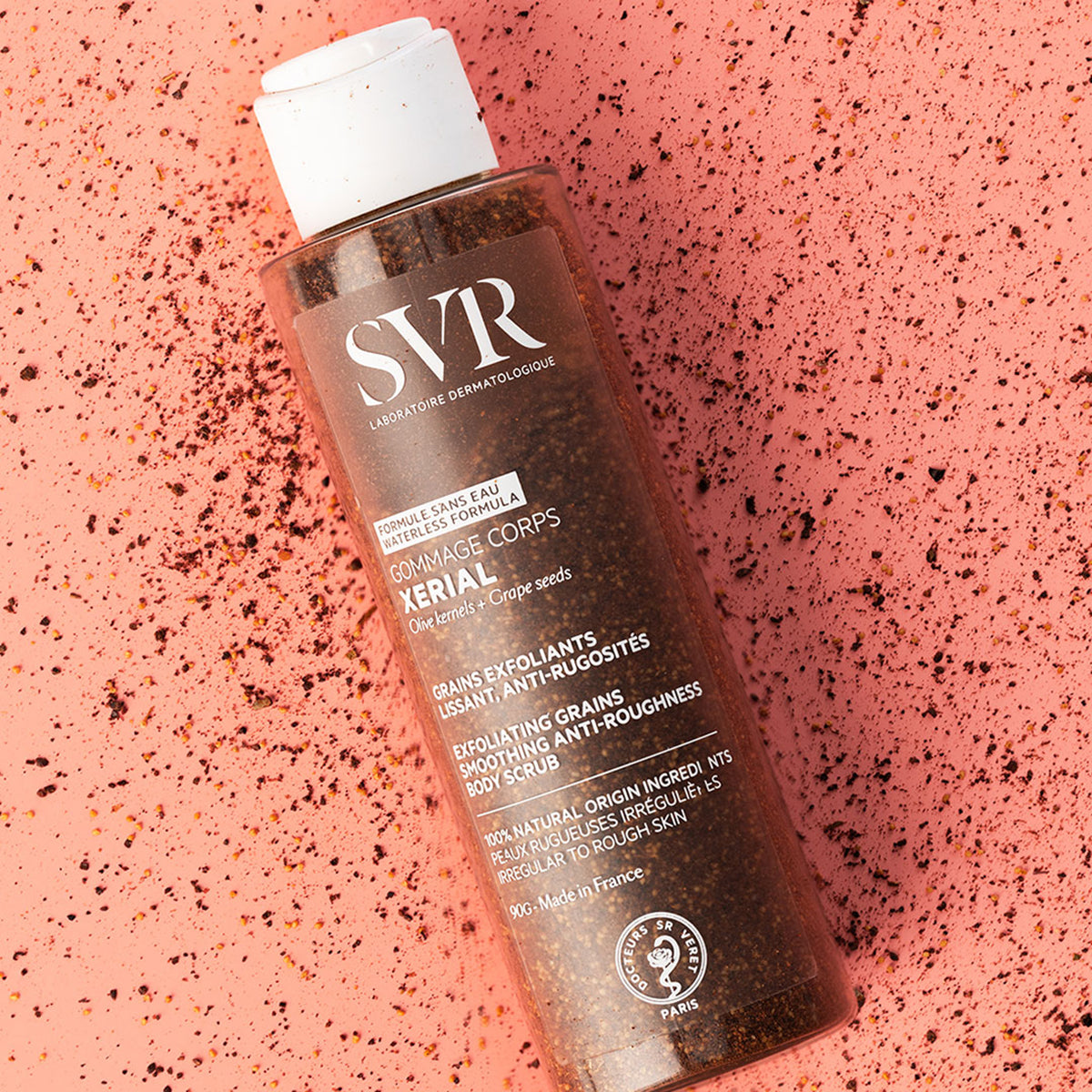 Envase cilíndrico de SVR Xerial Gommage con exfoliante granulado sobre un fondo texturizado de color coral.