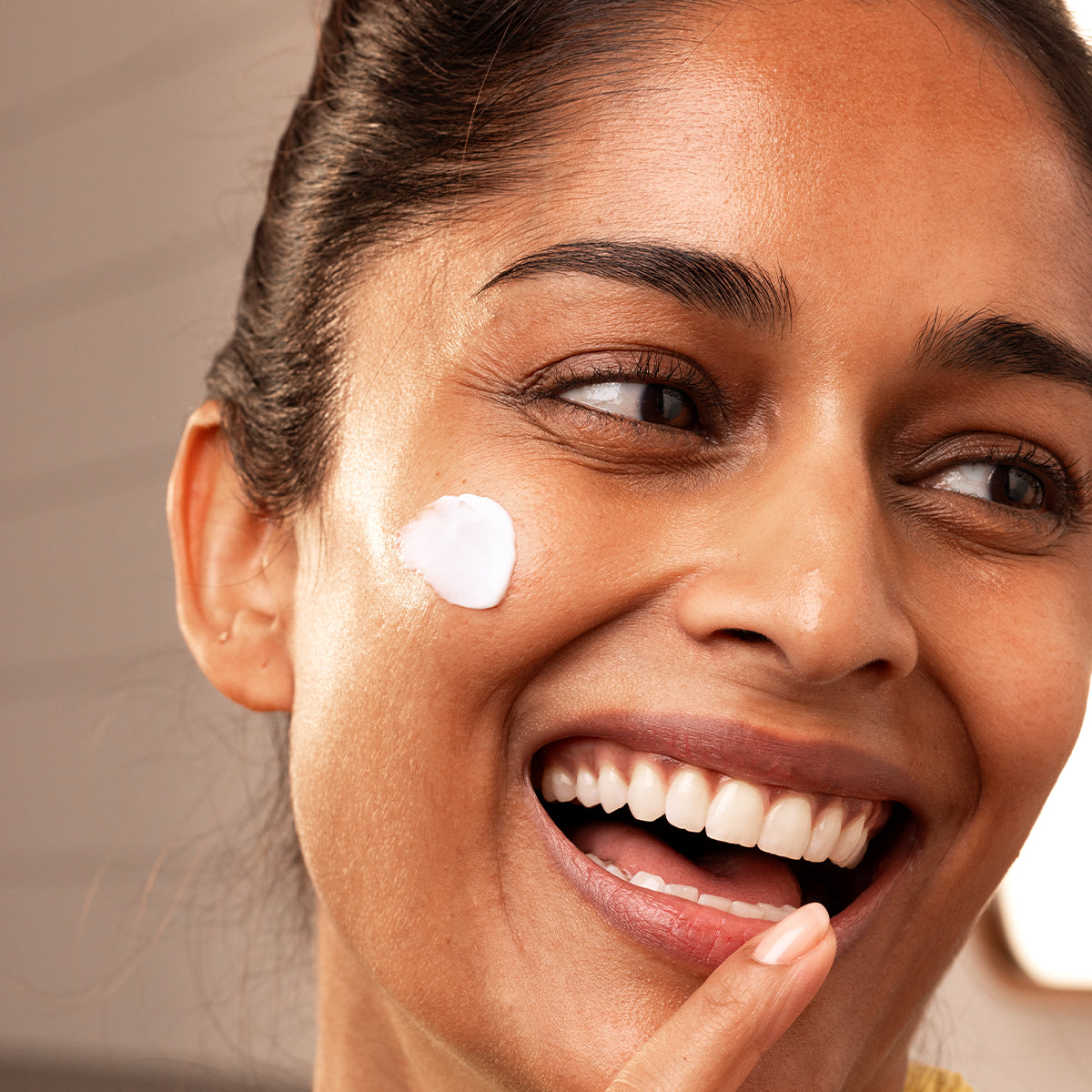 Mujer sonriente aplicándose una crema facial blanca de textura suave y regeneradora sobre la mejilla.