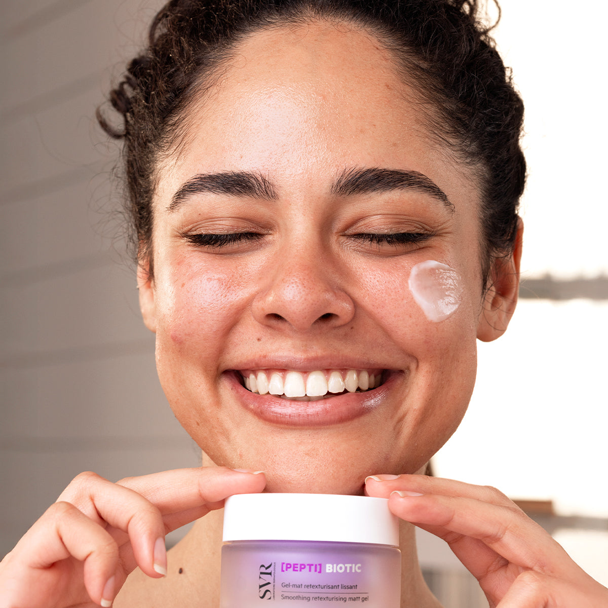 Mujer sonriente aplicando gel regenerador SVR [PEPTI] Biotic en su rostro con un toque de crema blanca en la mejilla.