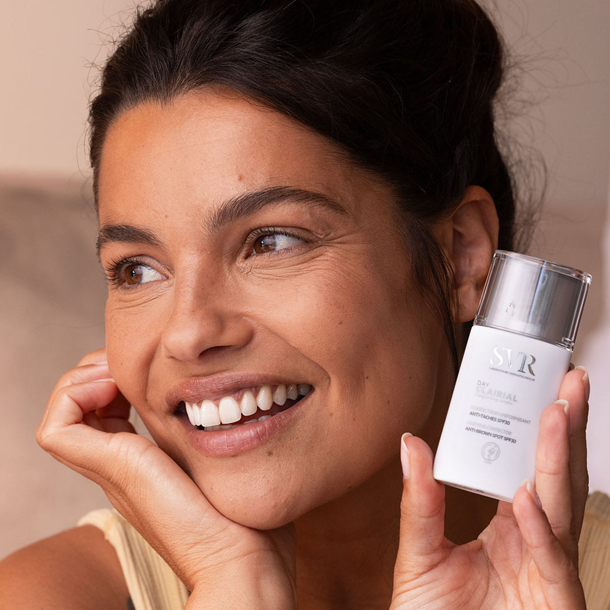 Mujer sonriente sosteniendo un envase blanco de SVR Clairial Day con tapa transparente y protección solar.
