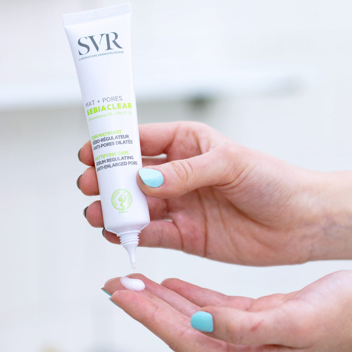 Tubo blanco de SVR Sebiaclear Mat+Pores dispensando una crema fluida sobre la palma de una mano con uñas azul claro.