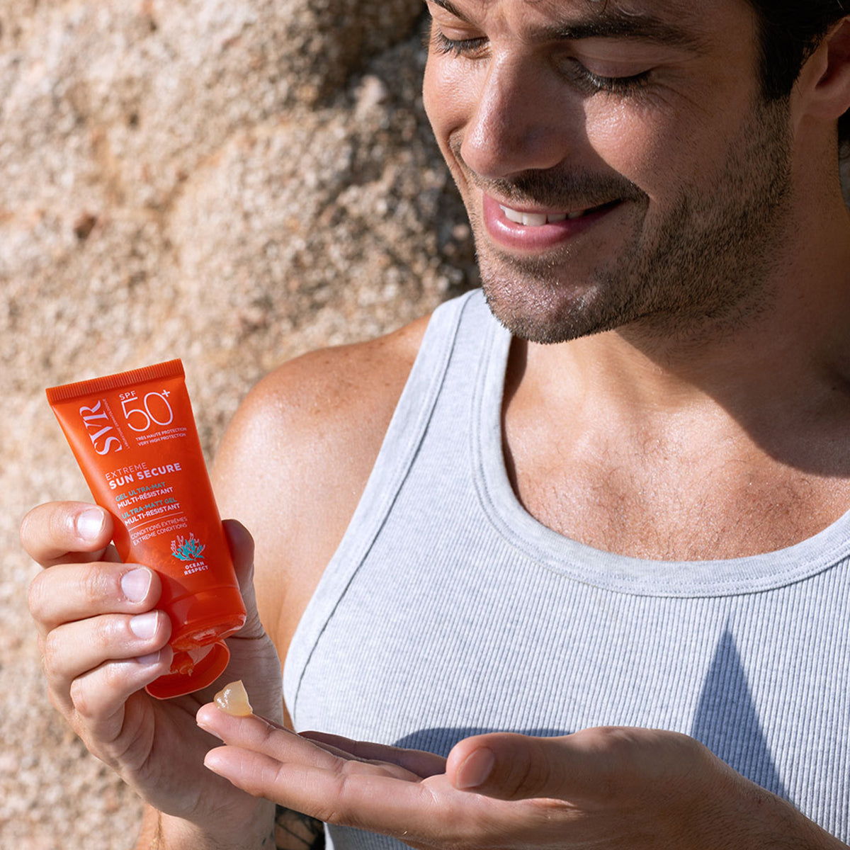 Hombre aplicando protector solar SVR Sun Secure Extrême en gel naranja sobre su mano frente a una pared de piedra.