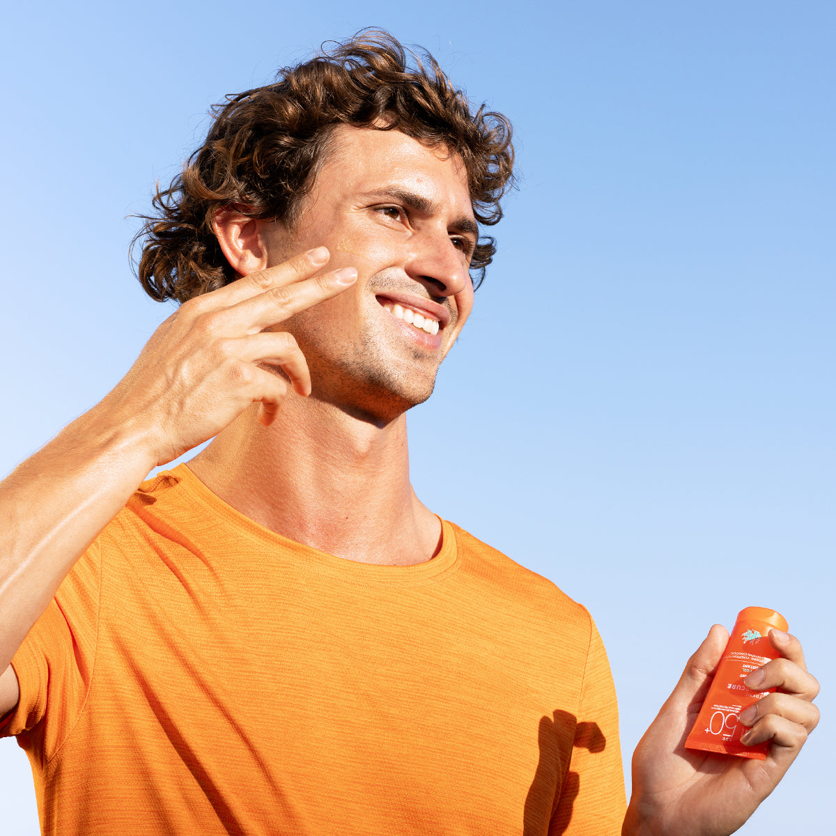 Hombre con camiseta naranja aplicando protector solar SUN SECURE Extreme SPF50+ en su rostro bajo un cielo azul despejado.
