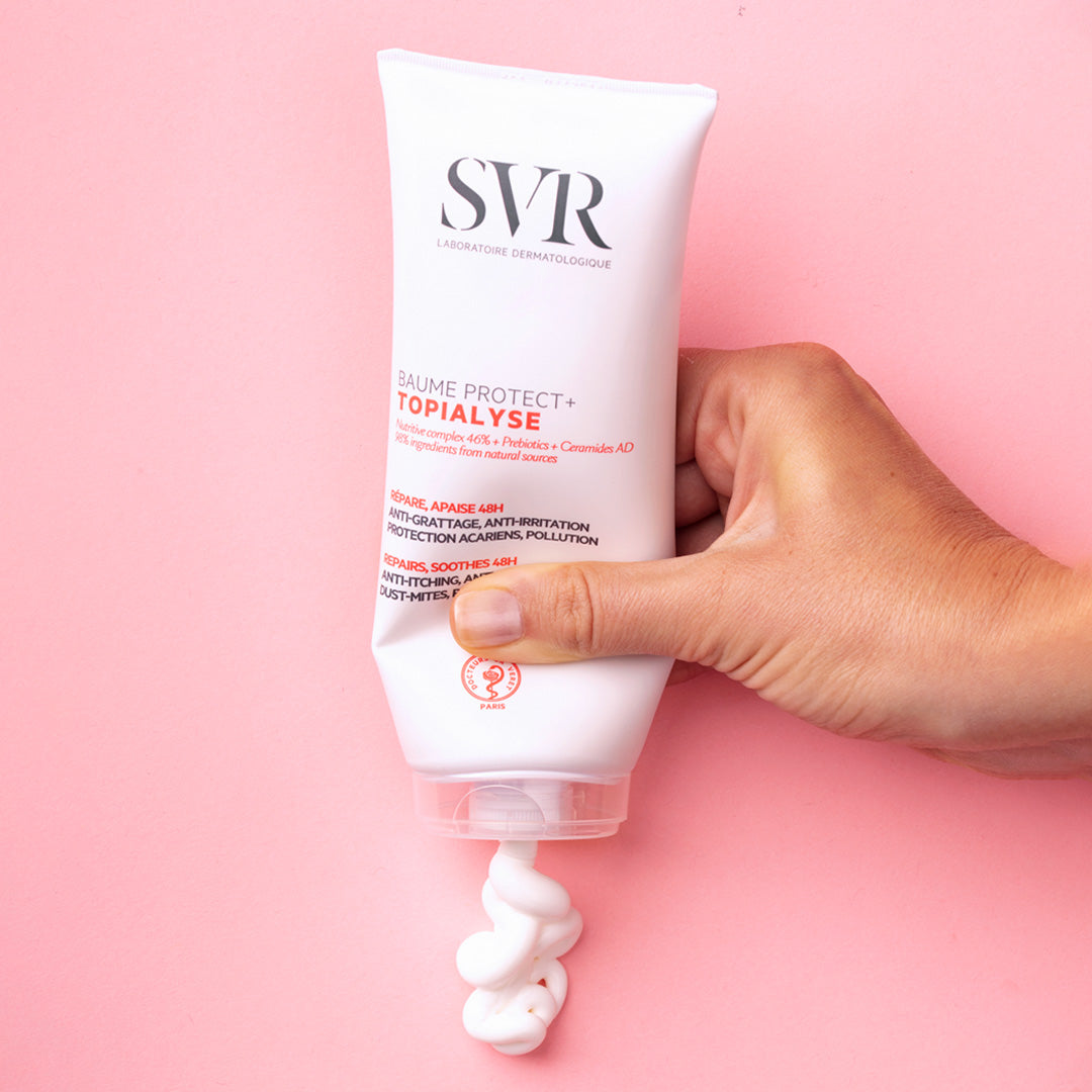 Mano apretando un tubo blanco de bálsamo SVR Topialyse Protect+ sobre un fondo rosa, mostrando la textura cremosa del producto.