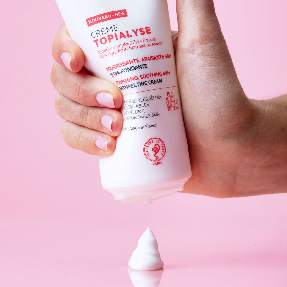 Mano sosteniendo un tubo blanco de crema Topialyse dispensando una gota de textura suave sobre un fondo rosa.
