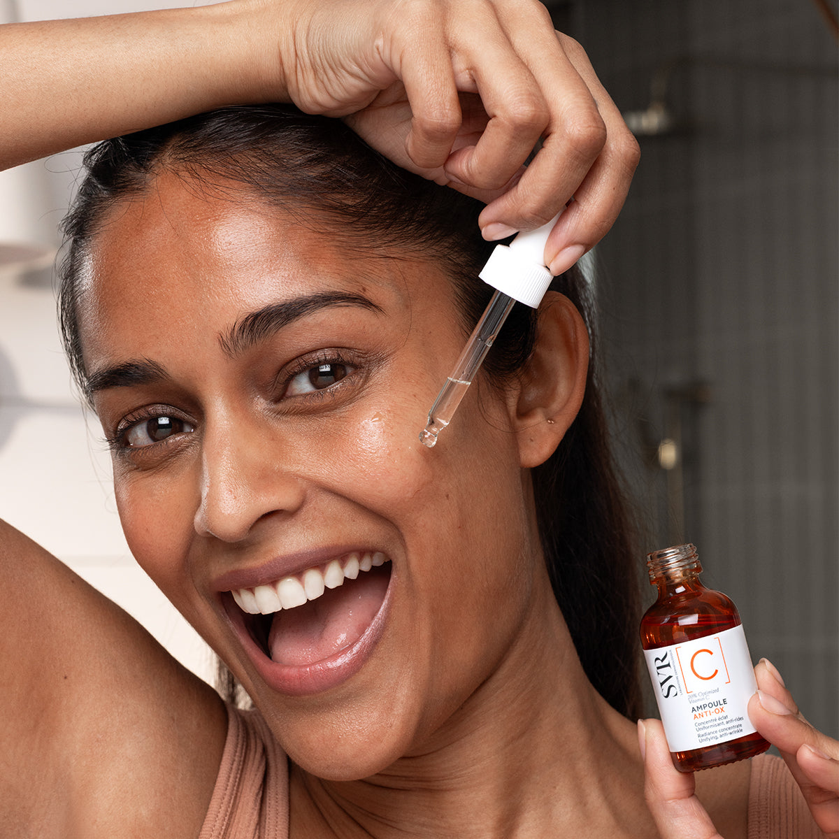 Mujer sonriente aplicando serum SVR Ampoule Anti-Ox con gotero sobre su rostro, frasco de vidrio ámbar con etiqueta blanca.