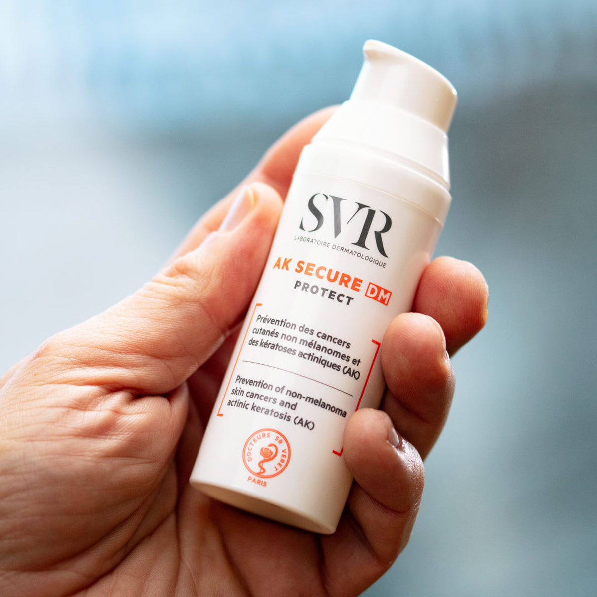 Frasco blanco de SVR AK SECURE DM Protect con dosificador, sostenido en una mano, para protección solar de pieles sensibles.