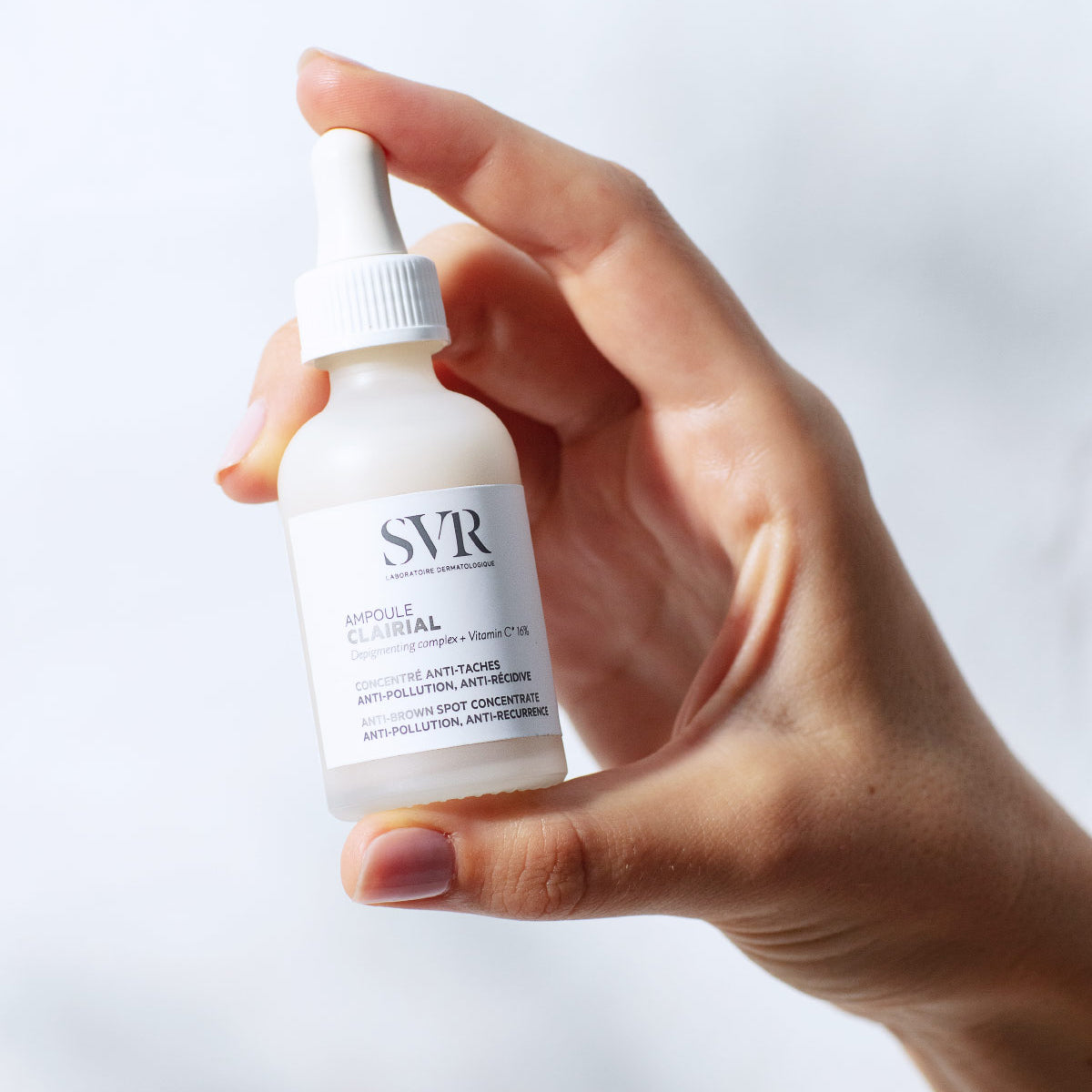 Frasco de vidrio esmerilado SVR Clairial Ampoule con gotero blanco sostenido por una mano sobre fondo claro.