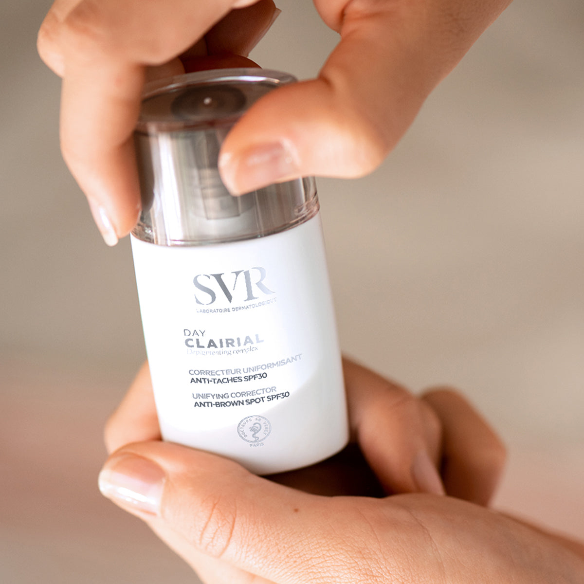 Manos sosteniendo un envase blanco cilíndrico de SVR Clairial Day, crema correctora antimanchas con protector solar SPF30.