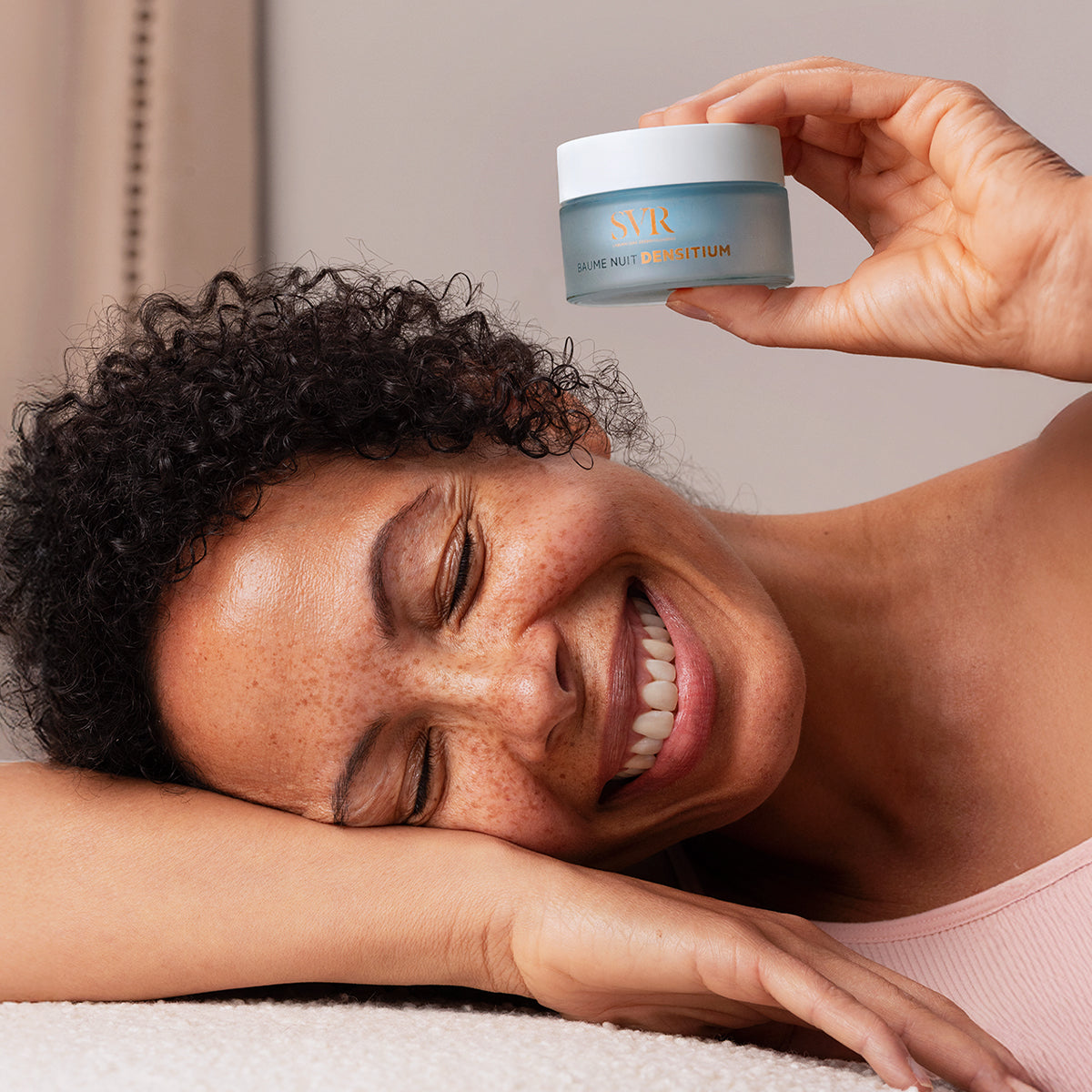 Smiling woman holding a frosted blue SVR DENSITIUM Baume Nuit jar with a white lid against a soft neutral background.