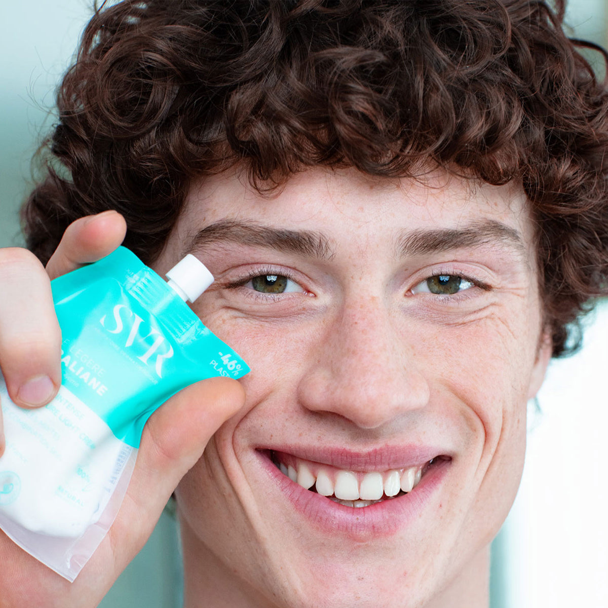 Joven sonriente sosteniendo el envase flexible y reciclable de color turquesa de la crema hidratante SVR Hydraliane Légere.