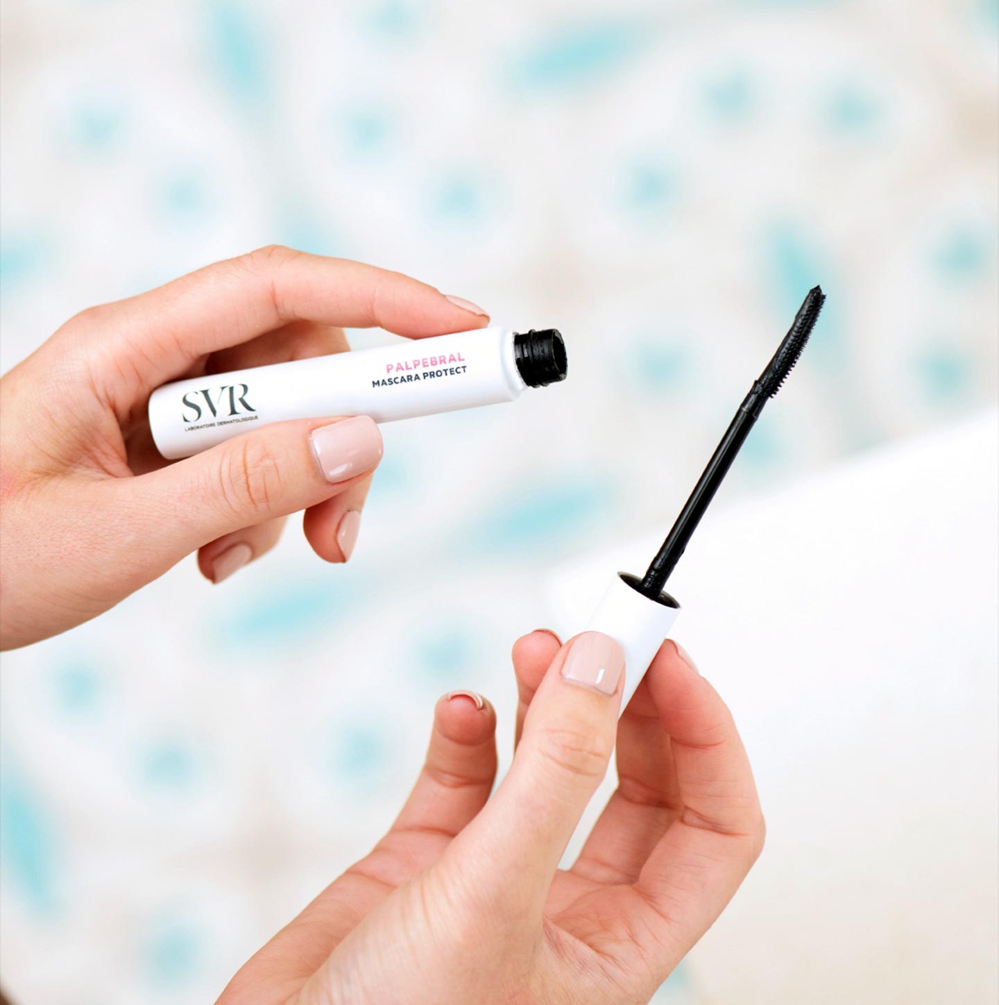 Manos sosteniendo el envase blanco y el aplicador negro de la máscara de pestañas SVR Palpebral Mascara Protect.