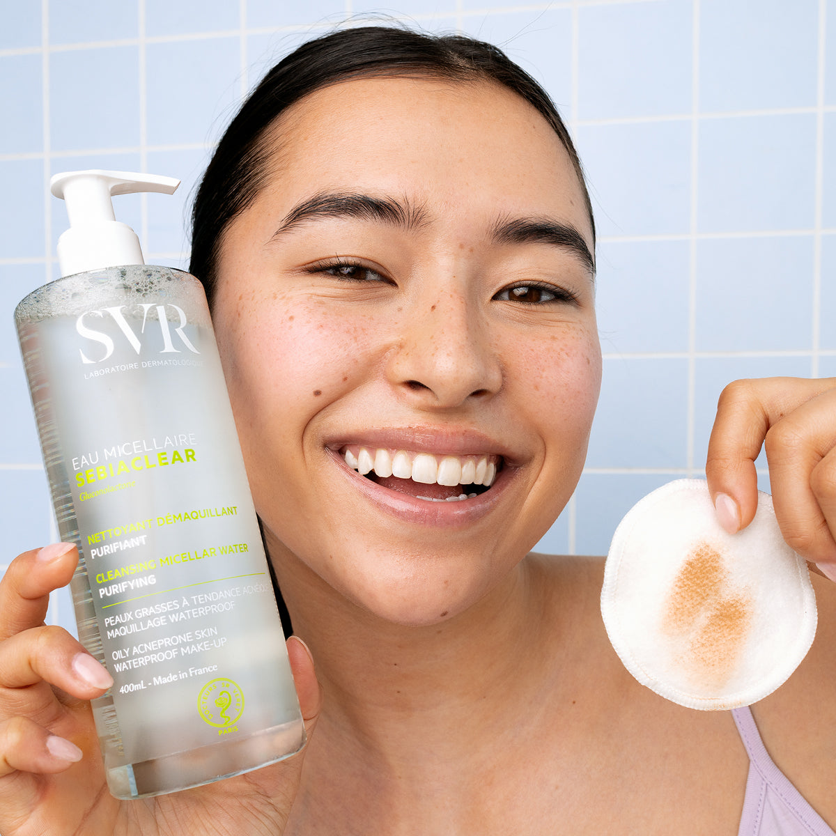 Mujer sonriente sosteniendo botella de 400ml de SVR Sebiaclear Eau Micellaire y un disco de algodón con restos de maquillaje.