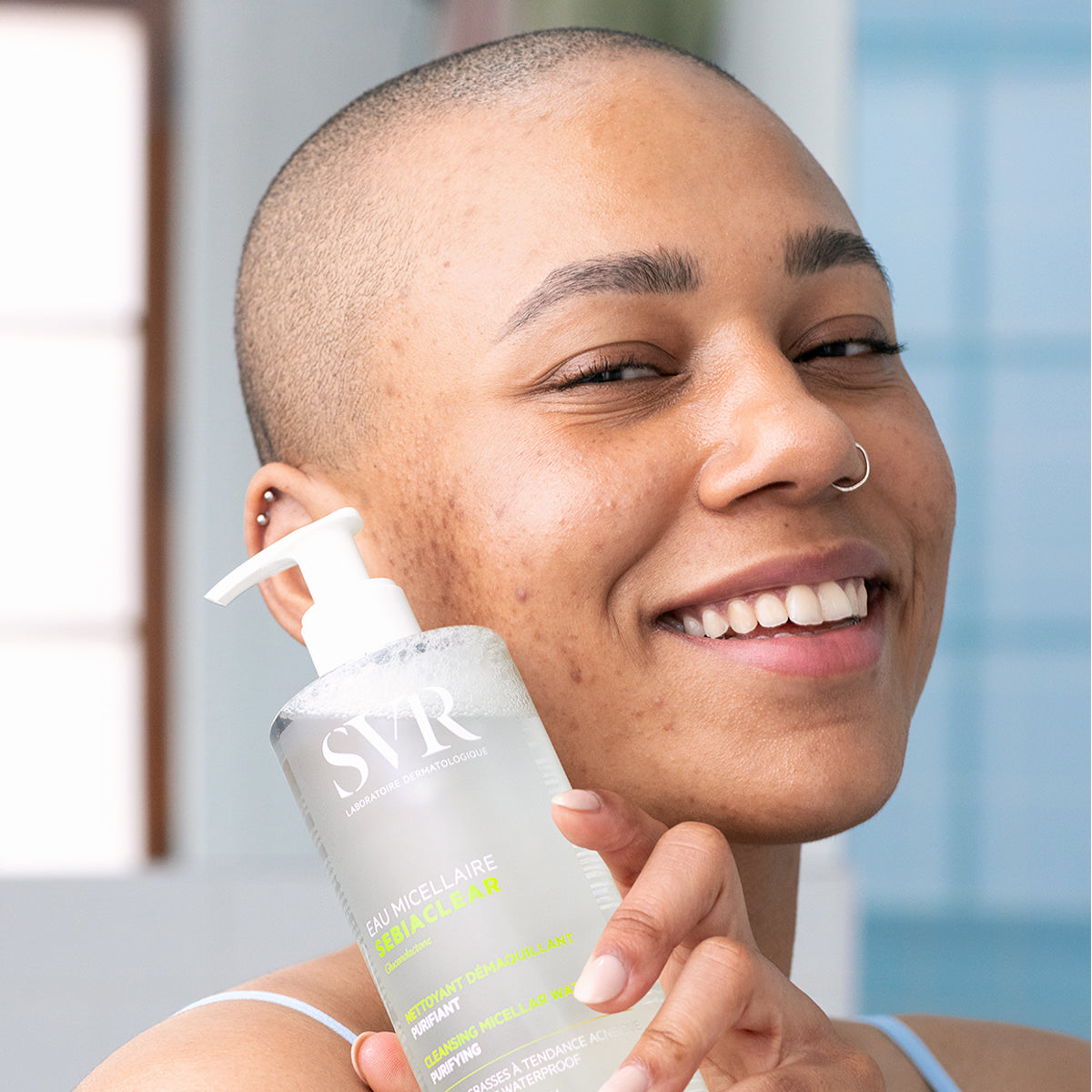 Mujer sonriente sosteniendo un frasco transparente con dosificador de agua micelar SVR Sebiaclear para pieles con tendencia acneica.