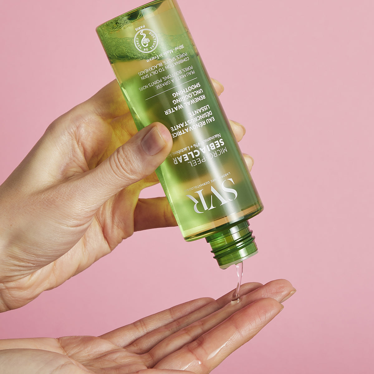 Frasco verde transparente de SVR Sebiaclear Micro-Peel vertiendo esencia líquida sobre una mano contra fondo rosa.