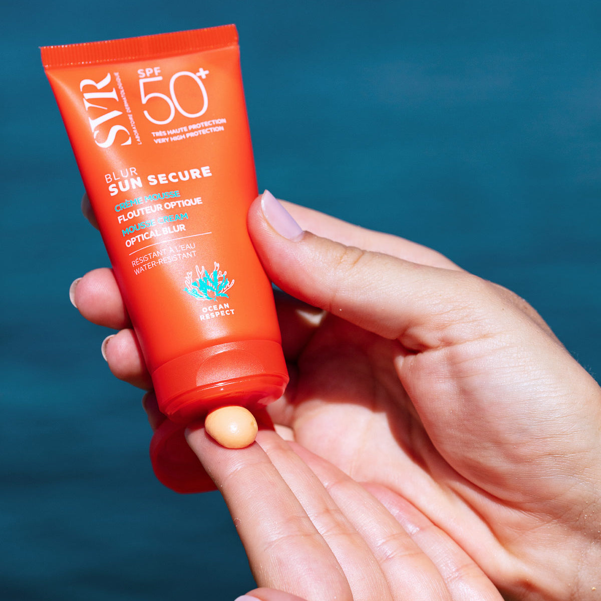 Tubo naranja de protector solar SVR Sun Secure SPF50+ con textura mousse albaricoque sobre una mano, fondo azul marino.