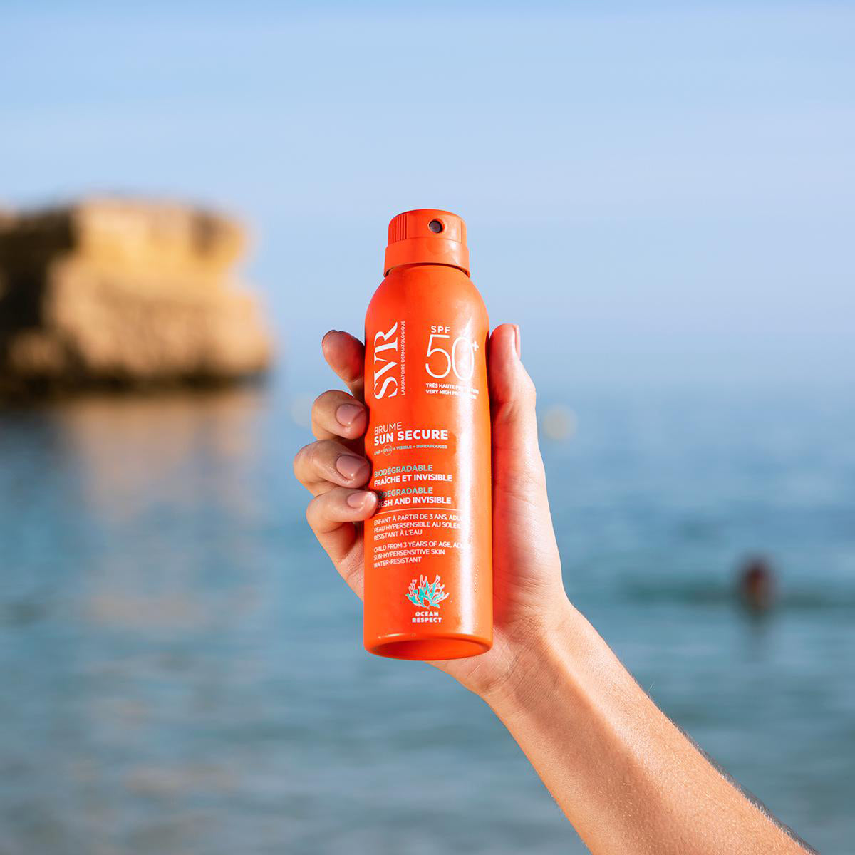 Mano sosteniendo un spray solar naranja SVR SUN SECURE Brume SPF50 frente a un fondo de playa desenfocado.