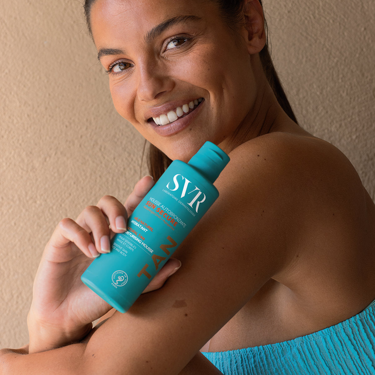 Mujer sonriente sosteniendo un frasco azul de mousse autobronceadora SVR Sun Secure para piel sensible.