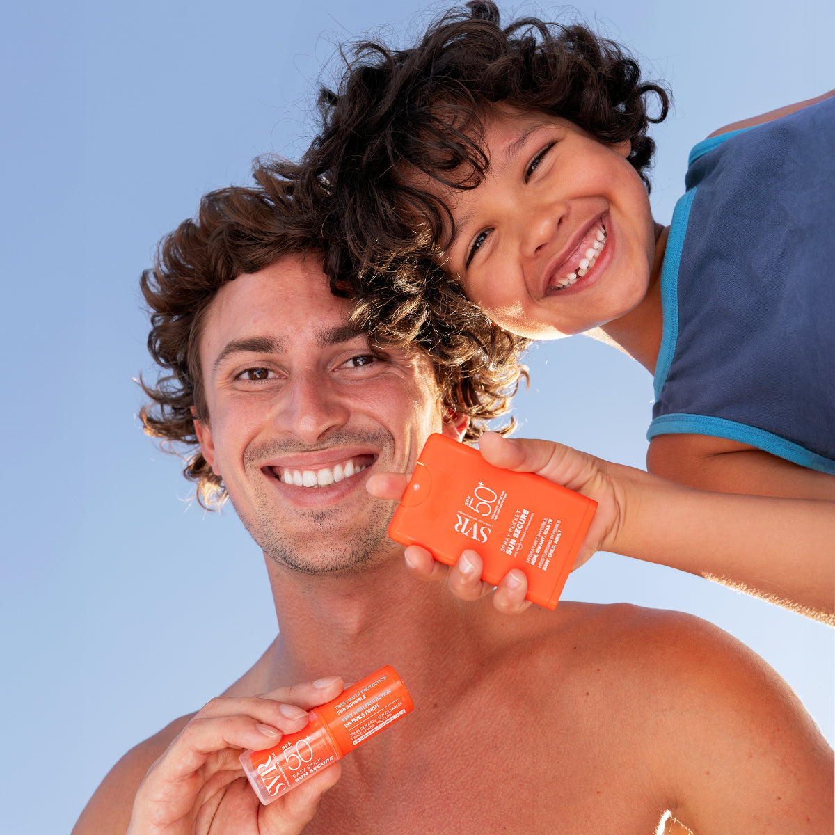 Padre e hijo sonrientes sosteniendo un protector solar naranja SVR Sun Secure SPF50+ contra un cielo azul despejado.