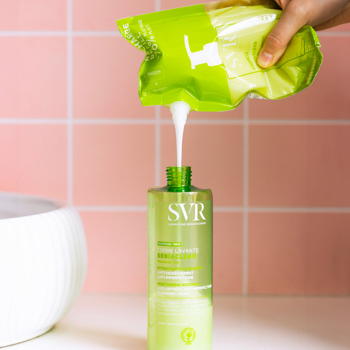 Recarga de SVR Sebiaclear Creme Lavante vertiendo la crema limpiadora blanca en un envase verde de 400ml sobre fondo rosa.