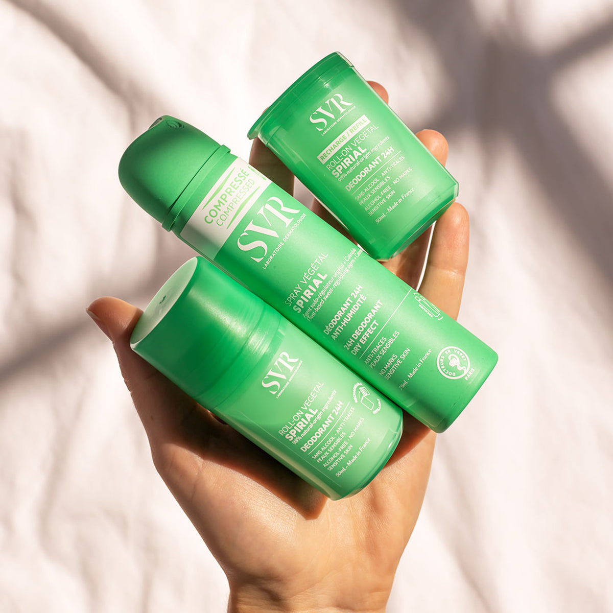 Mano sosteniendo tres desodorantes verdes SVR Spirial: un spray comprimido y dos envases roll-on sobre fondo blanco.