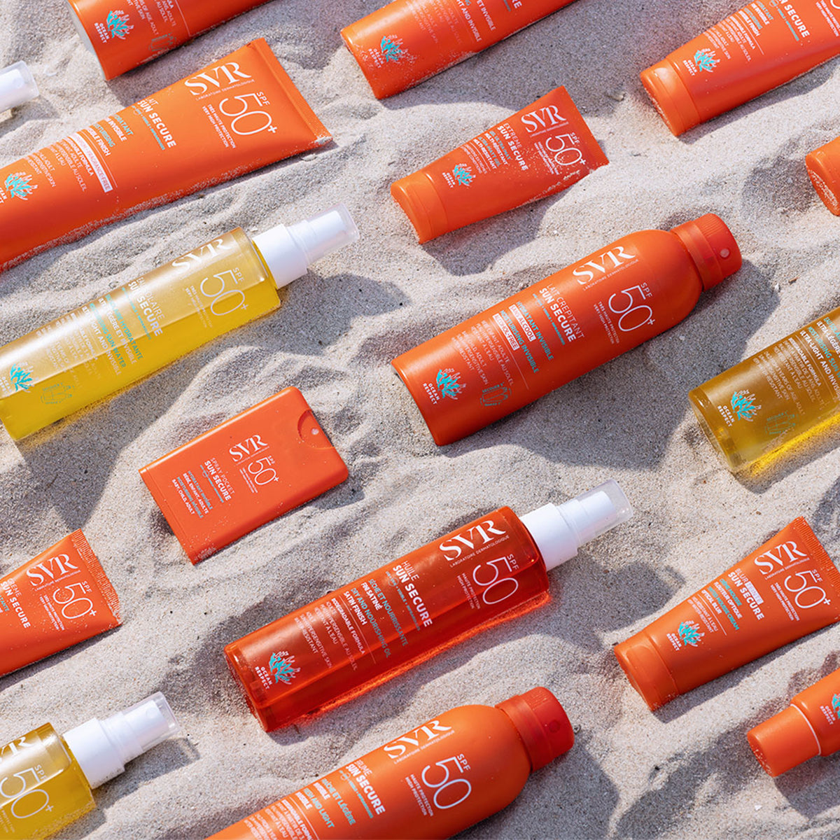 Botellas y tubos de protector solar SVR Sun Secure SPF50+ en tonos naranja y amarillo dispuestos sobre arena de playa.