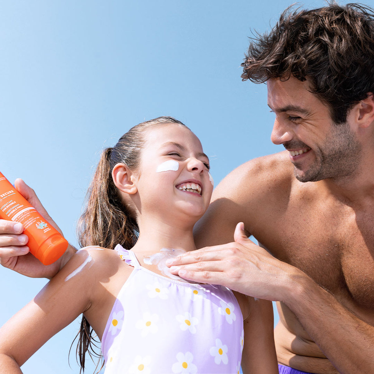 Father applying orange SUN SECURE SPF50+ sunscreen lotion onto a smiling girl's shoulder against a blue sky.