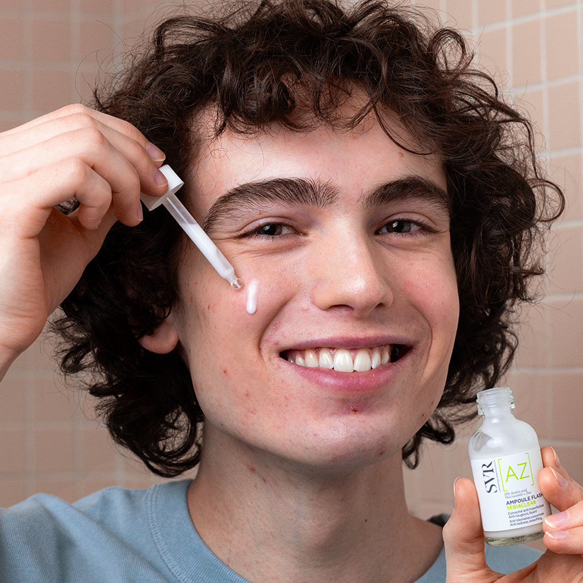 Joven aplicando el sérum SVR Sebiaclear Ampoule Flash con gotero sobre su rostro frente a un fondo de azulejos rosados.