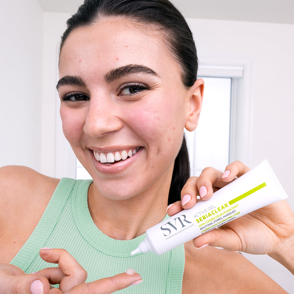 Mujer sonriente sosteniendo el tubo blanco de SVR Sebiaclear Active Gel con una gota de producto en el dedo.