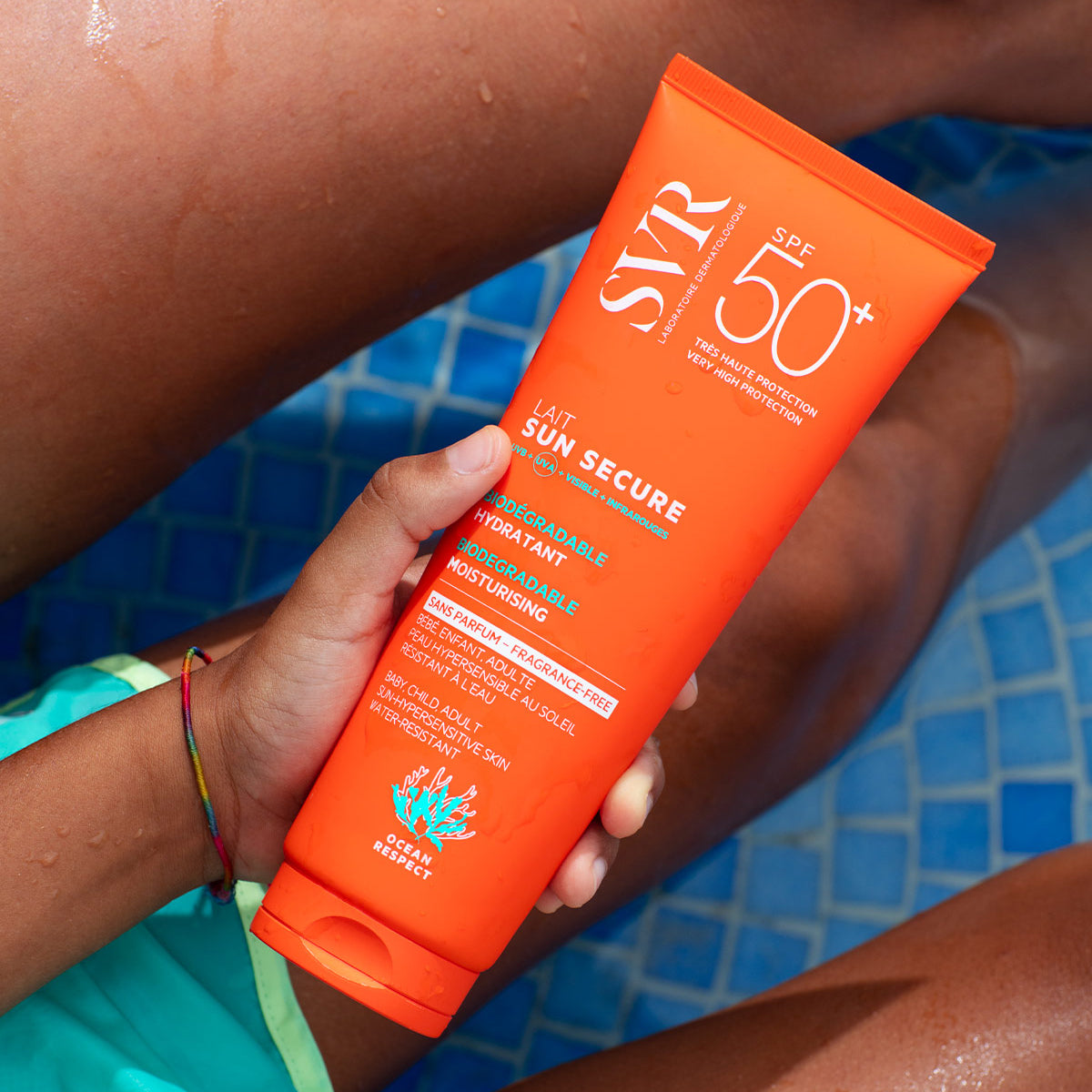 Bright orange SVR SUN SECURE SPF50+ sunscreen tube held by a person in a blue tiled swimming pool.