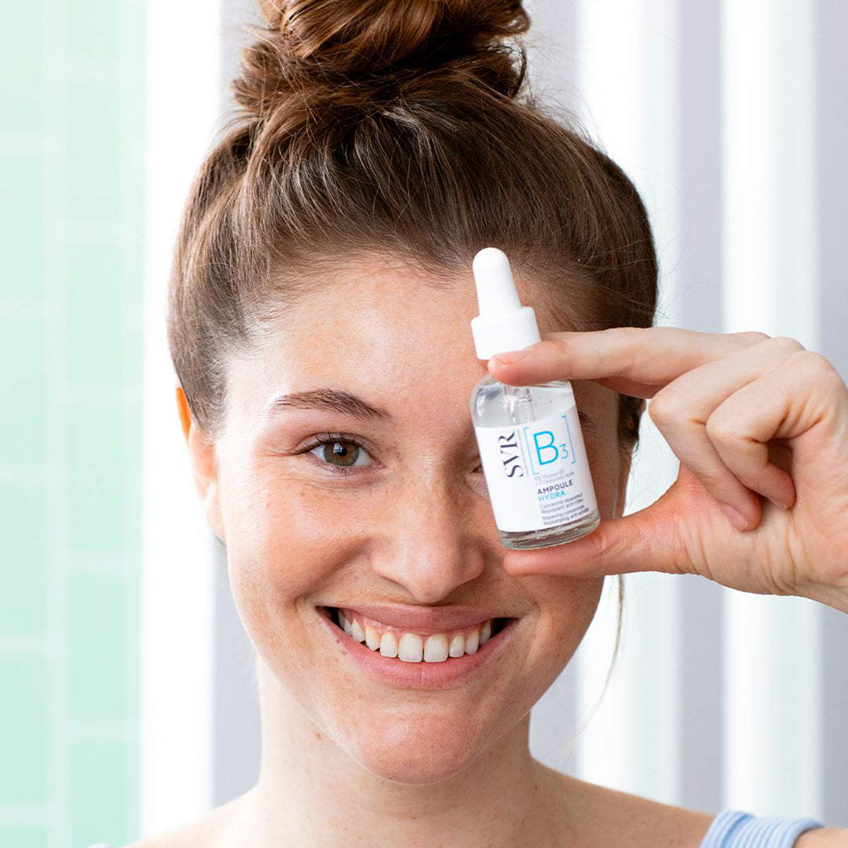 Mujer sonriente sosteniendo un frasco de vidrio con gotero de SVR Ampoule B3 Hydra frente a su rostro para el cuidado facial.