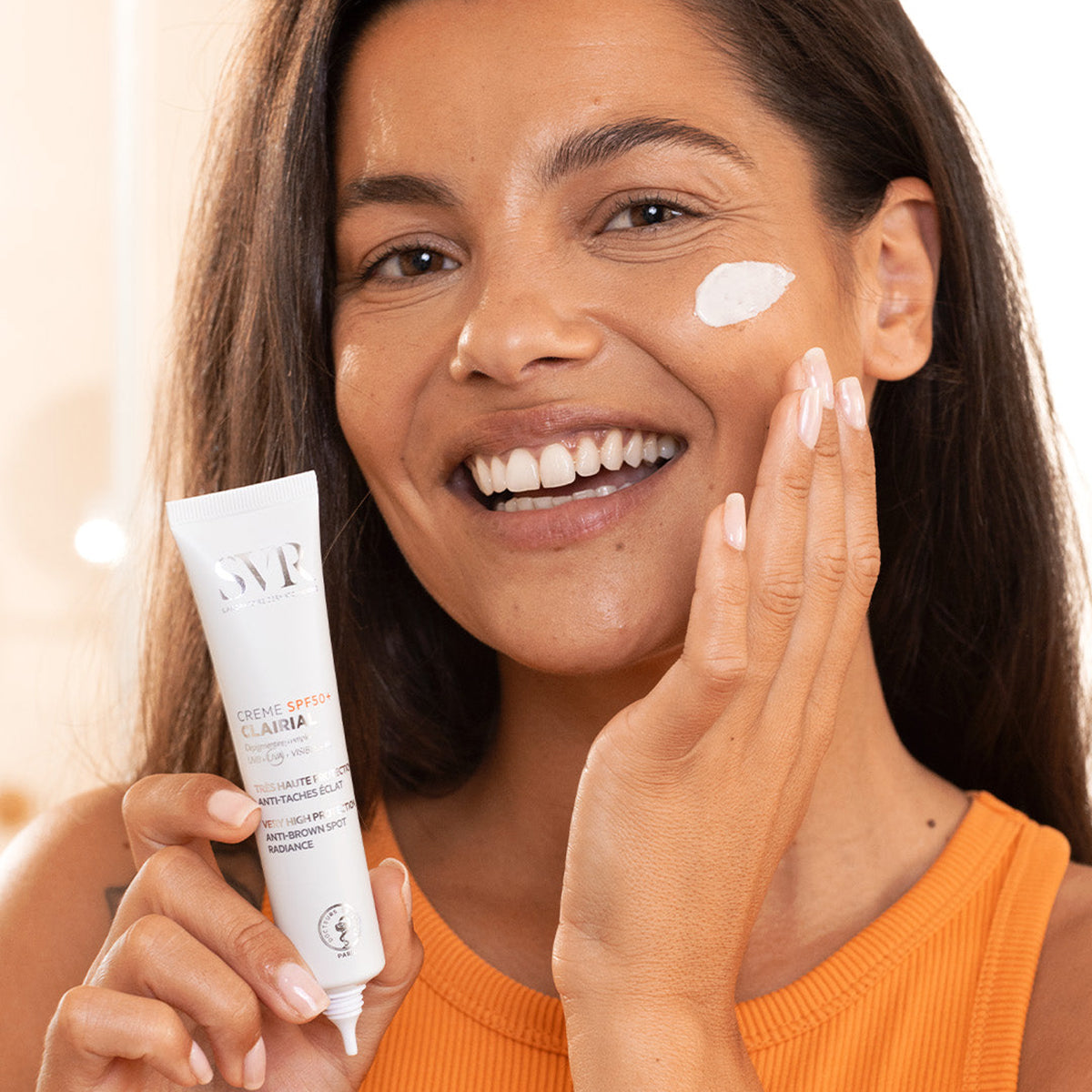 Smiling woman applying white SVR Clairial SPF50+ cream to her cheek from a white squeeze tube.