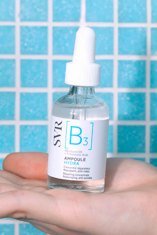 SVR Ampoule Hydra serum bottle with a white dropper cap held in a hand against a blue tiled background.