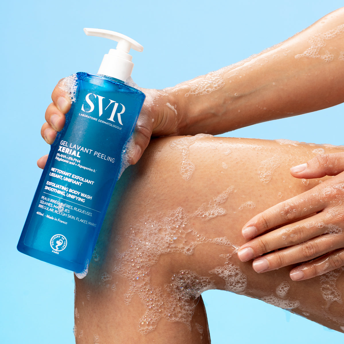 Envase azul de 400ml de gel exfoliante SVR Xerial con dosificador, aplicado sobre una pierna con espuma jabonosa.