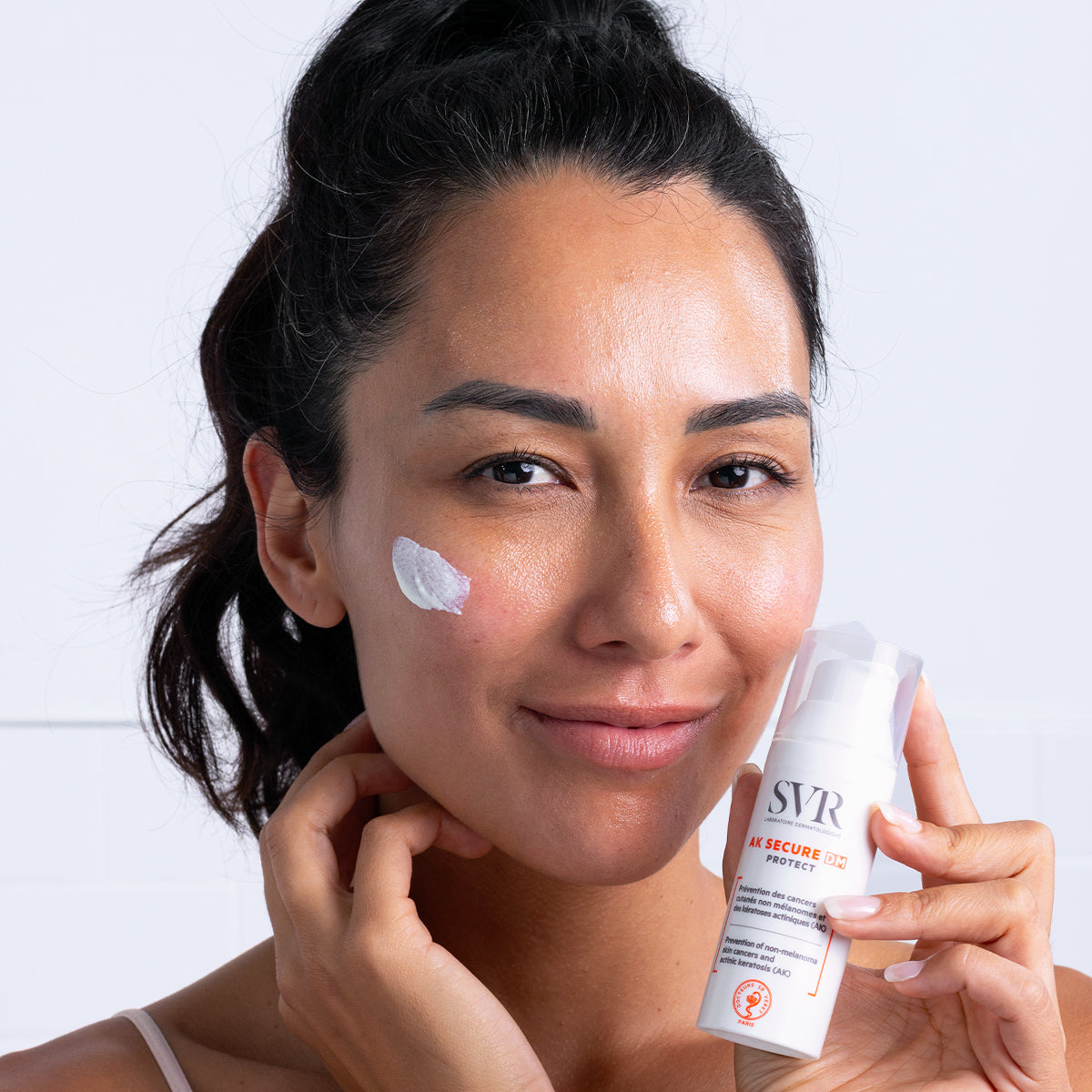 Mujer aplicando crema solar protectora SVR AK SECURE DM Protect en su mejilla, sosteniendo el envase blanco con dosificador.