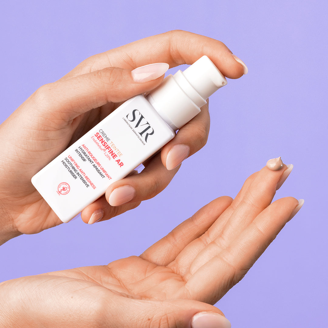 Manos sosteniendo un envase blanco de SVR Sensifine AR Creme Teinté con una gota de crema con color en el dedo.