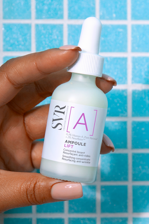 Frasco de vidrio SVR Ampoule [A] Lift con gotero blanco sostenido en la mano sobre un fondo de azulejos azules.