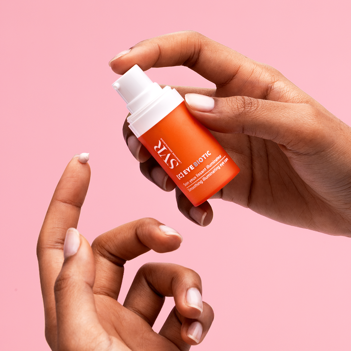 Manos sosteniendo un envase naranja de contorno de ojos SVR [C] EYE BIOTIC con una gota de crema en el dedo.
