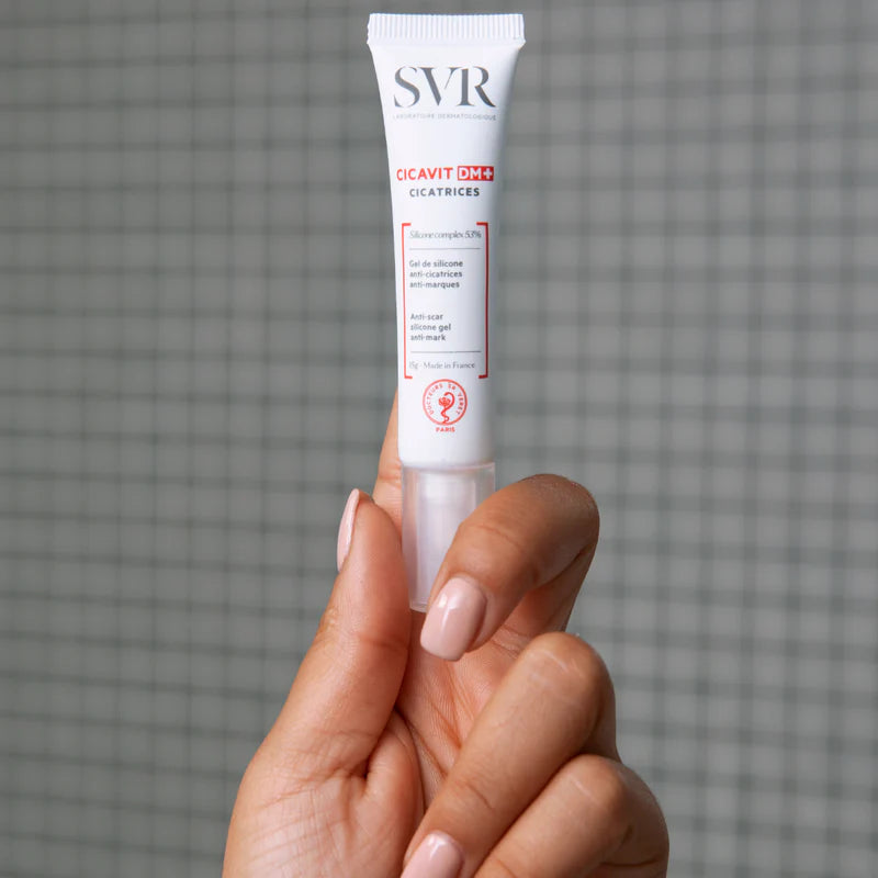 Tubo blanco de gel cicatrizante SVR Cicavit DM+ de 15g sostenido por una mano sobre fondo de azulejos grises.