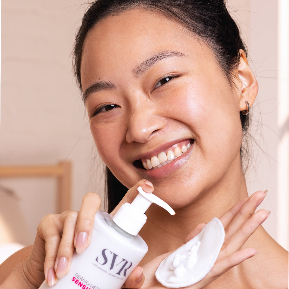 Mujer sonriente sosteniendo el limpiador SVR Sensifine en envase blanco con dosificador y un disco de algodón con crema.
