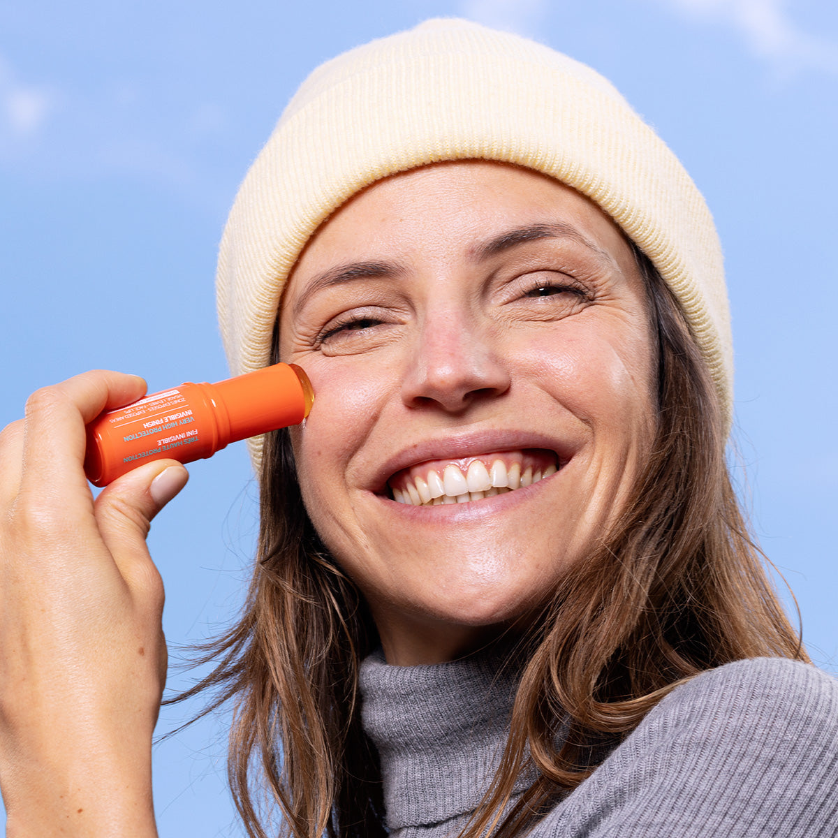Mujer sonriente aplicando protector solar en barra naranja SUN SECURE SPF50+ sobre su mejilla contra un fondo azul.