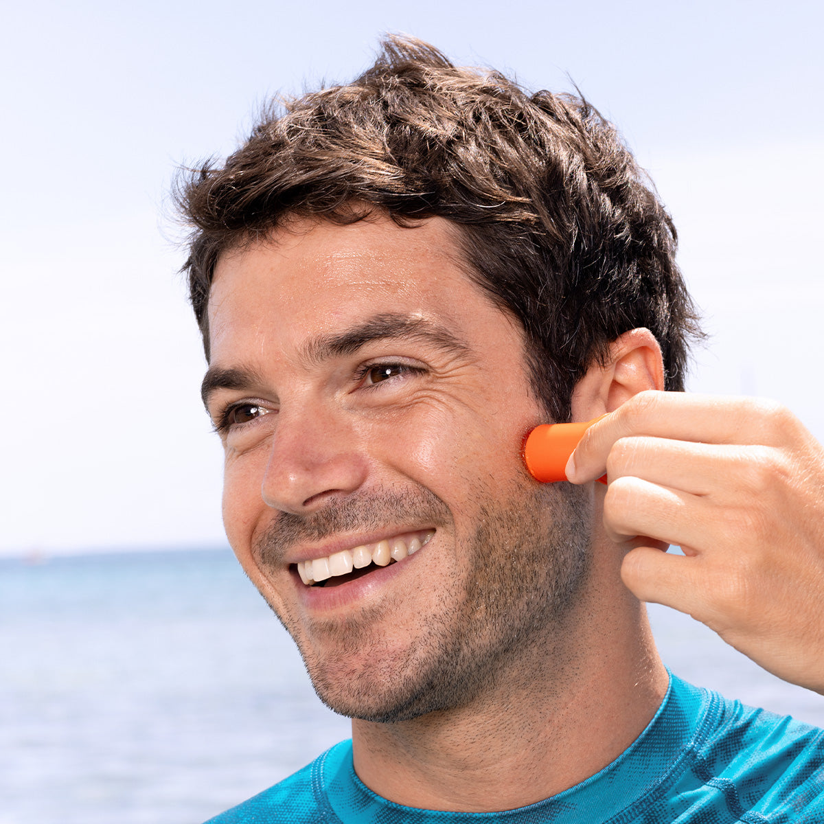 Hombre sonriente aplicándose protector solar en barra naranja SUN SECURE SPF50+ en la mejilla frente al mar.