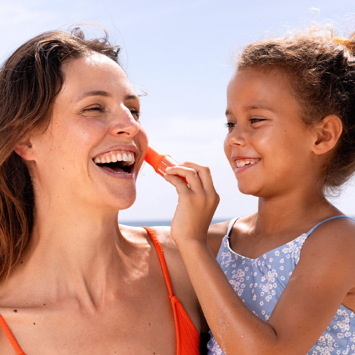 Niña aplicando protector solar en barra naranja SUN SECURE SPF50+ en el rostro de su madre sonriente al aire libre.