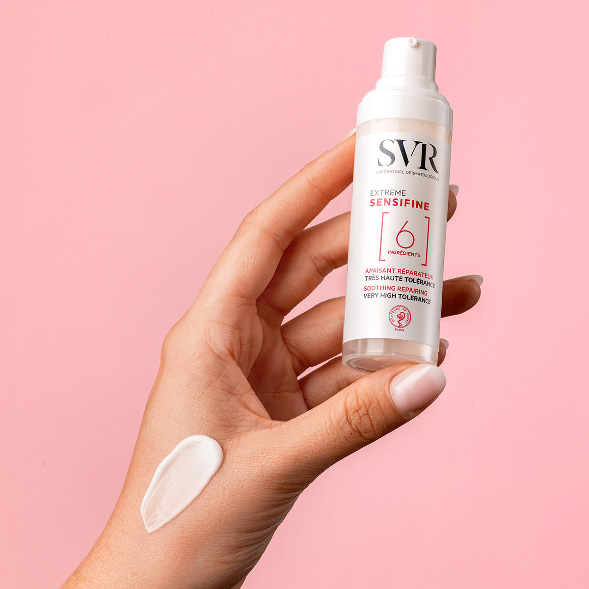 Mano sosteniendo envase blanco de SVR Sensifine Extrême con una muestra de crema blanca sobre la piel y fondo rosa.