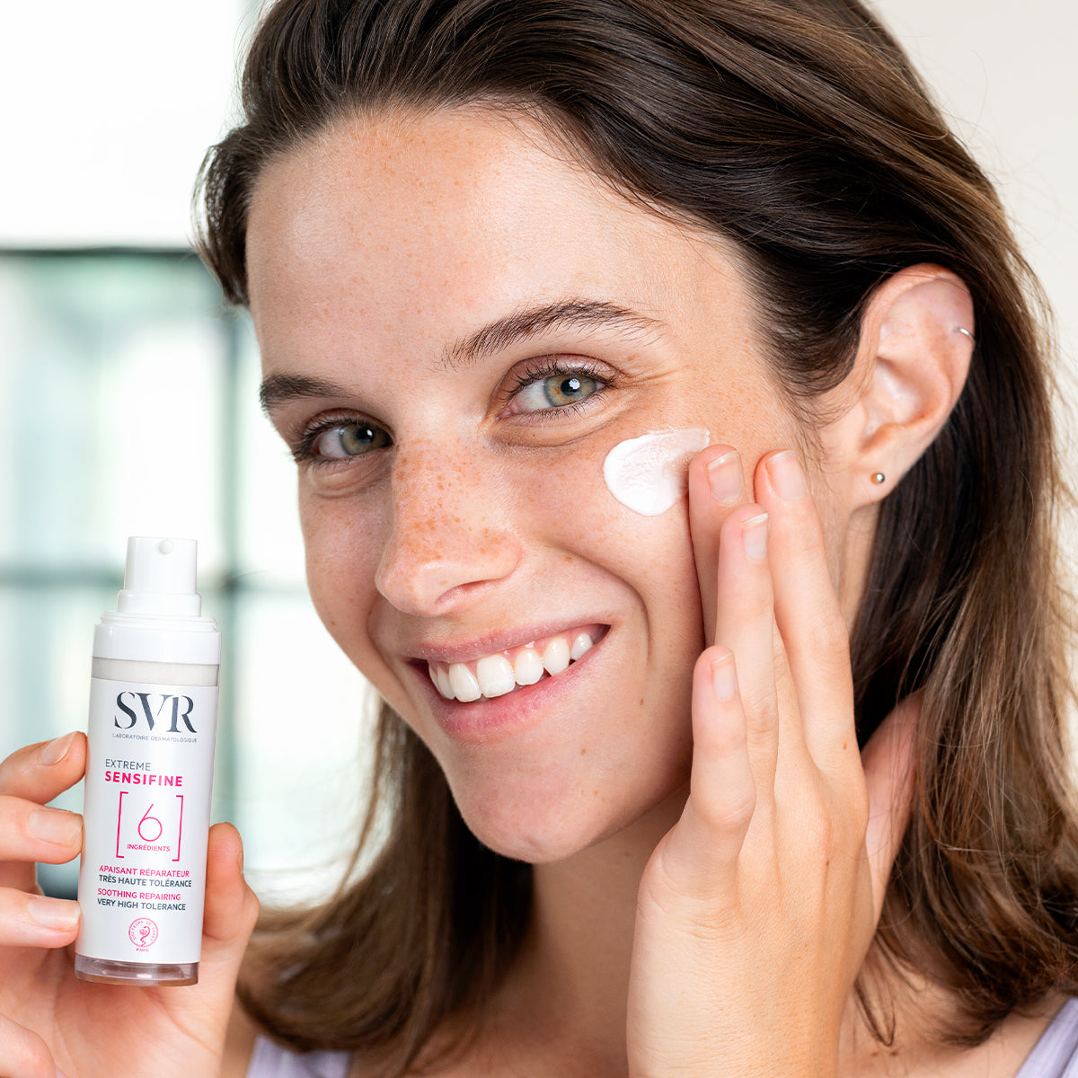 Mujer sonriente aplicando crema blanca SVR SENSIFINE Extrême desde un envase dosificador minimalista sobre su mejilla.