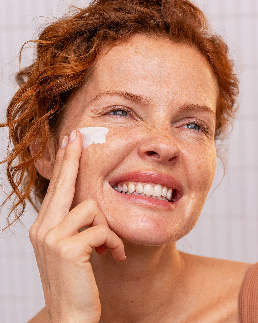 Smiling woman with red hair applying white SUN SECURE SPF50+ sunscreen cream to her cheek.