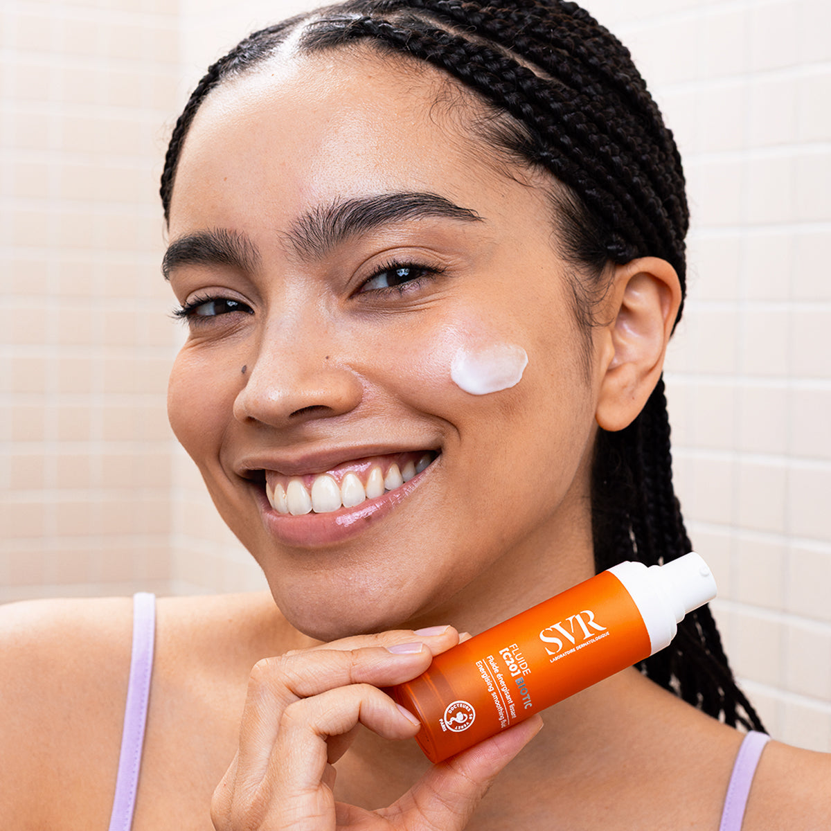 Mujer sonriente aplicando Fluide [C20] BIOTIC de SVR, un frasco naranja con crema blanca sobre su mejilla para una piel luminosa.