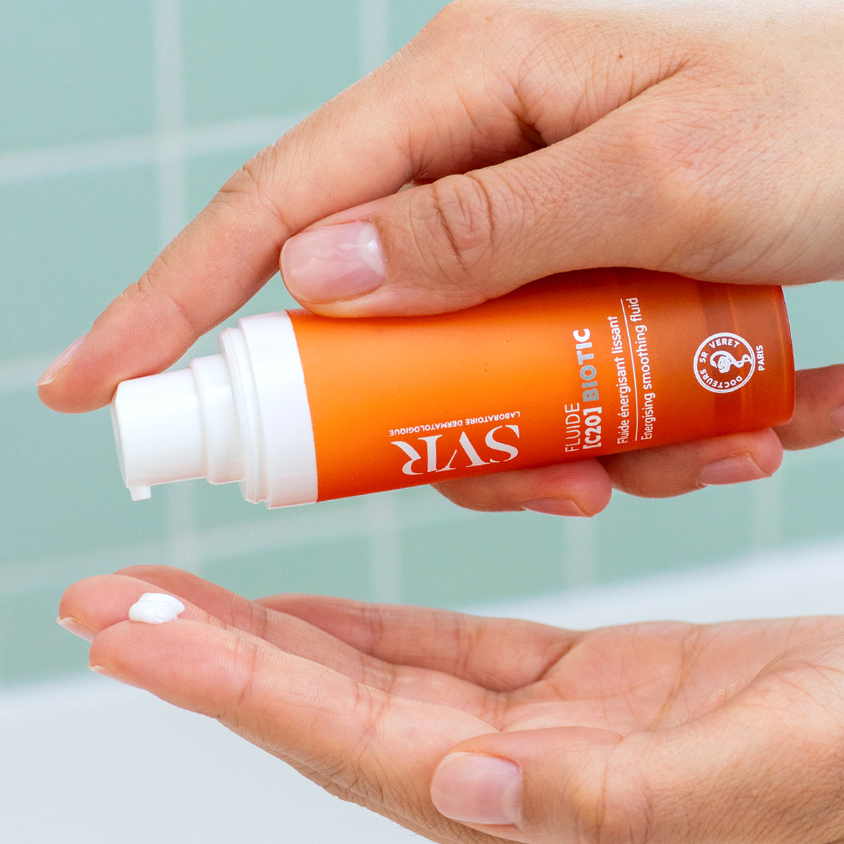 Frasco naranja de SVR Fluide [C20] BIOTIC dispensando una gota de crema blanca sobre la palma de una mano.
